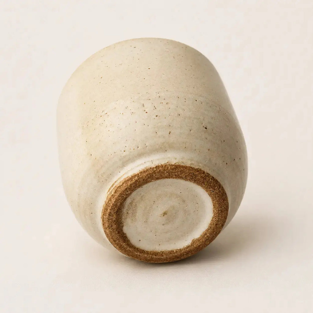 A handmade ceramic espresso cup with a rounded, slightly tapered shape, featuring a speckled off-white glaze and a contrasting brown clay rim at the base, photographed on a neutral light beige background.