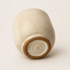 A handmade ceramic espresso cup with a rounded, slightly tapered shape, featuring a speckled off-white glaze and a contrasting brown clay rim at the base, photographed on a neutral light beige background.
