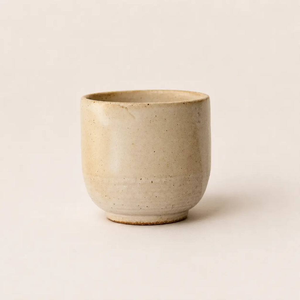 A small handmade ceramic espresso cup with a textured, speckled beige glaze, photographed against a solid, warm off-white background.