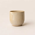 A small handmade ceramic espresso cup with a textured, speckled beige glaze, photographed against a solid, warm off-white background.