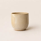 A small handmade ceramic espresso cup with a textured, speckled beige glaze, photographed against a solid, warm off-white background.