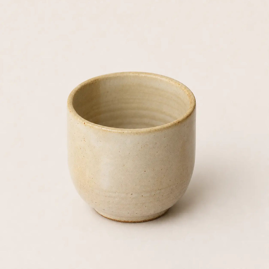 A small handmade ceramic espresso cup with a textured, speckled beige glaze, photographed against a solid, warm off-white background.