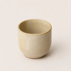 A small handmade ceramic espresso cup with a textured, speckled beige glaze, photographed against a solid, warm off-white background.