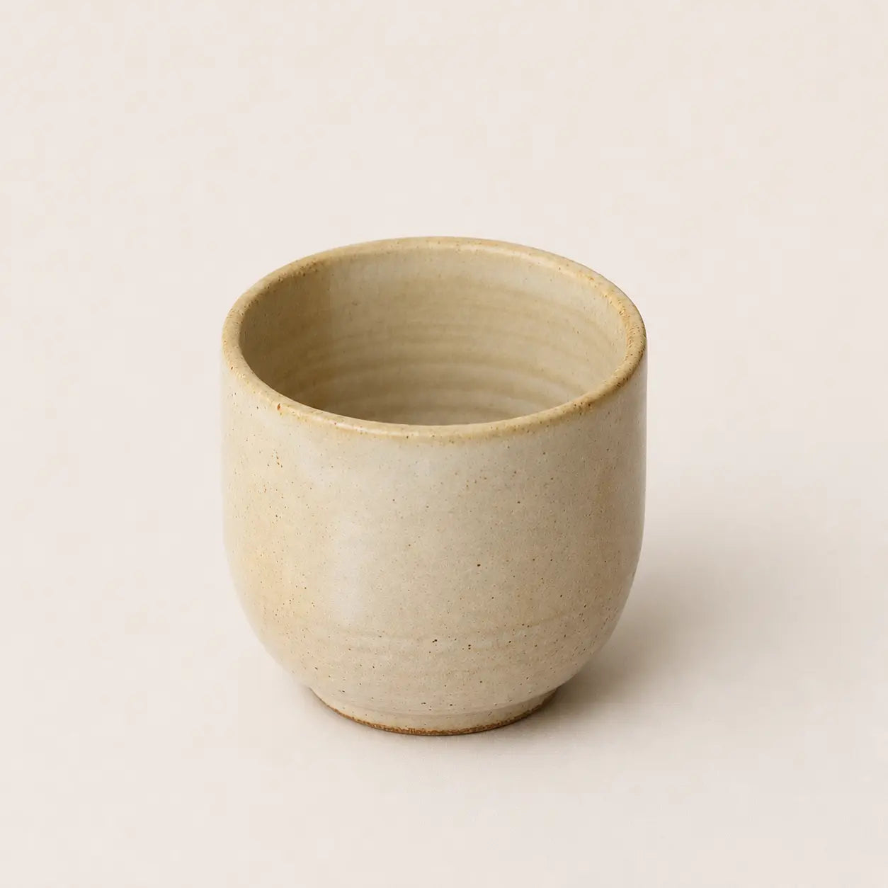 A small handmade ceramic espresso cup with a textured, speckled beige glaze, photographed against a solid, warm off-white background.