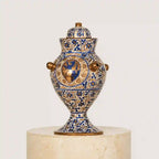 An ornate blue and gold decorative urn with a lid, displayed on a light beige stone pedestal against a plain off-white background.