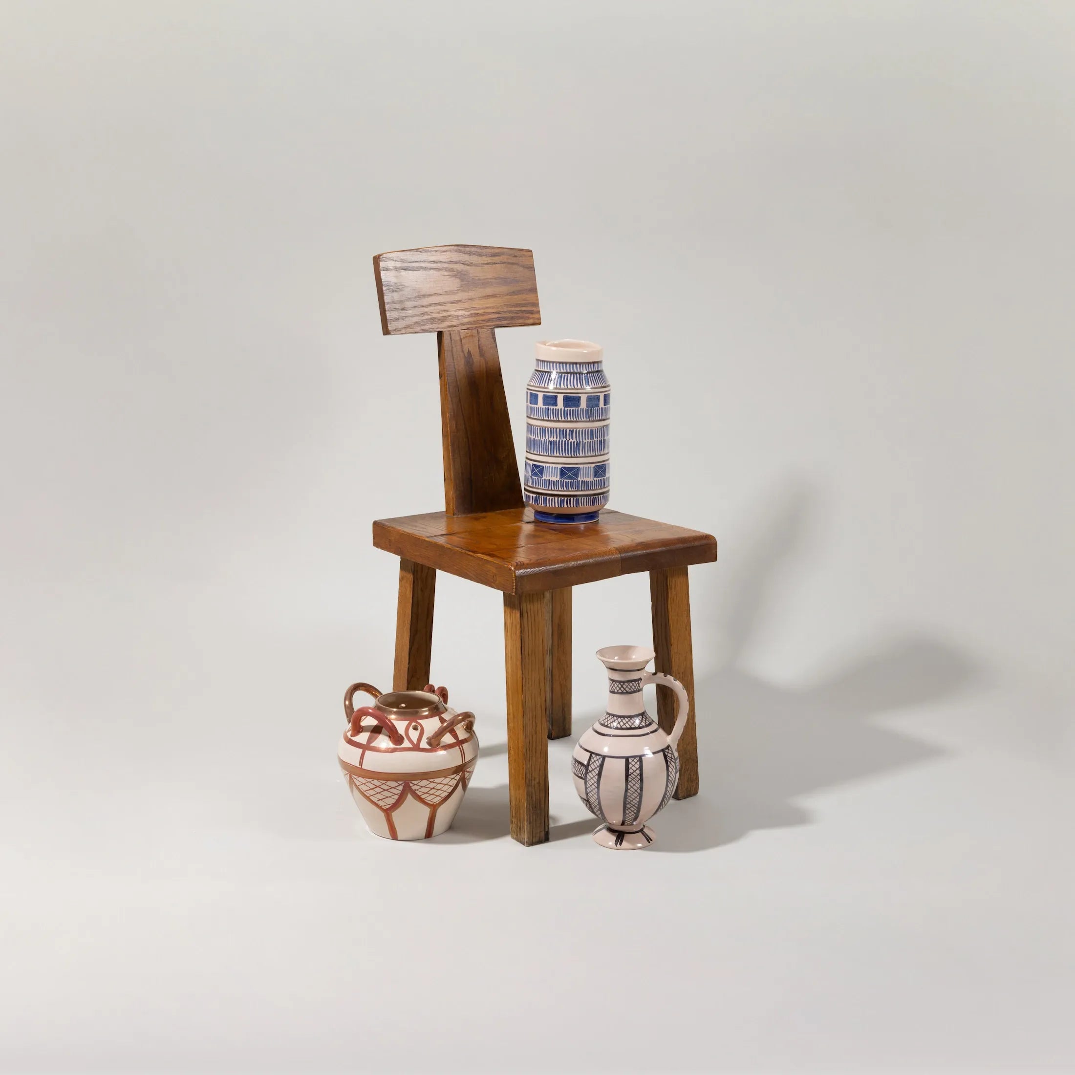 Handcrafted ceramic vessels with geometric painted patterns arranged beside a wooden chair in a minimalist studio setting.