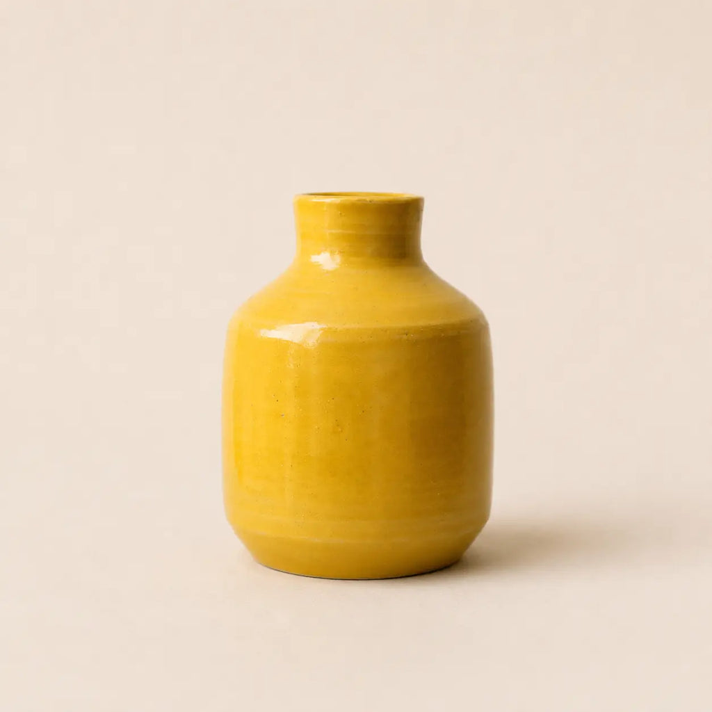 A small glossy ceramic vase in a warm mustard yellow color, centered against a solid, light beige background.