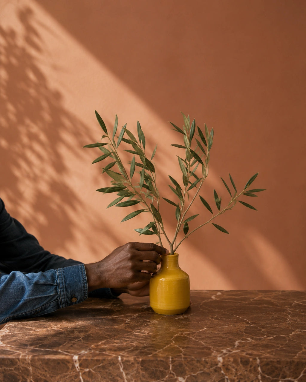A small yellow ceramic vase holding green olive branches, placed on a brown marble table against a terracotta wall with soft leaf shadows.