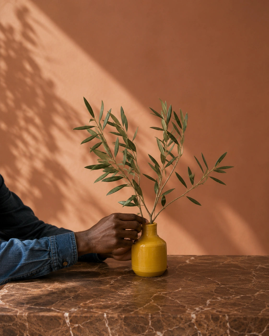 A small yellow ceramic vase holding green olive branches, placed on a brown marble table against a terracotta wall with soft leaf shadows.