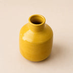 A small glossy ceramic vase with a rounded body and short neck, finished in a warm mustard yellow glaze, set against a plain light beige background.