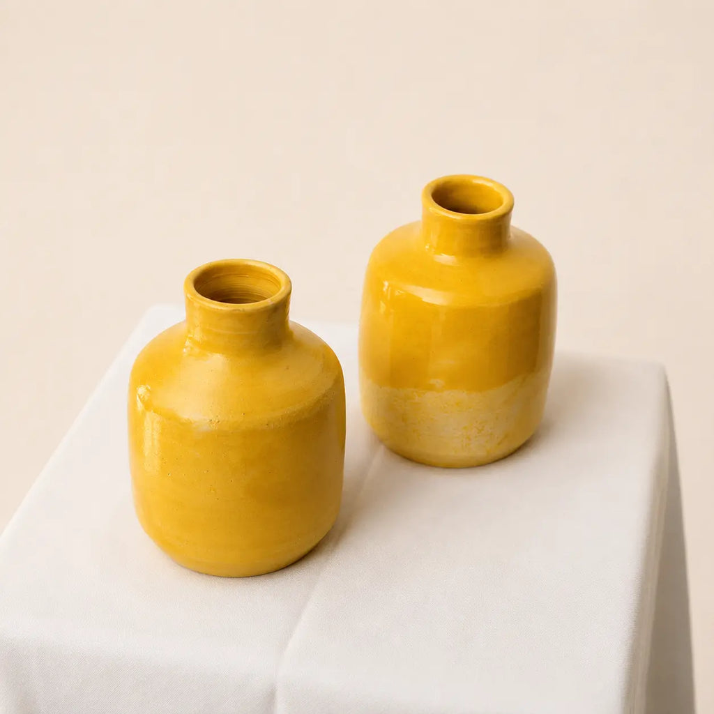 Two small glossy yellow ceramic bud vases with short necks, displayed on a white fabric-covered pedestal against a warm beige background.