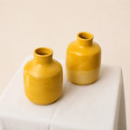 Two small glossy yellow ceramic bud vases with short necks, displayed on a white fabric-covered pedestal against a warm beige background.