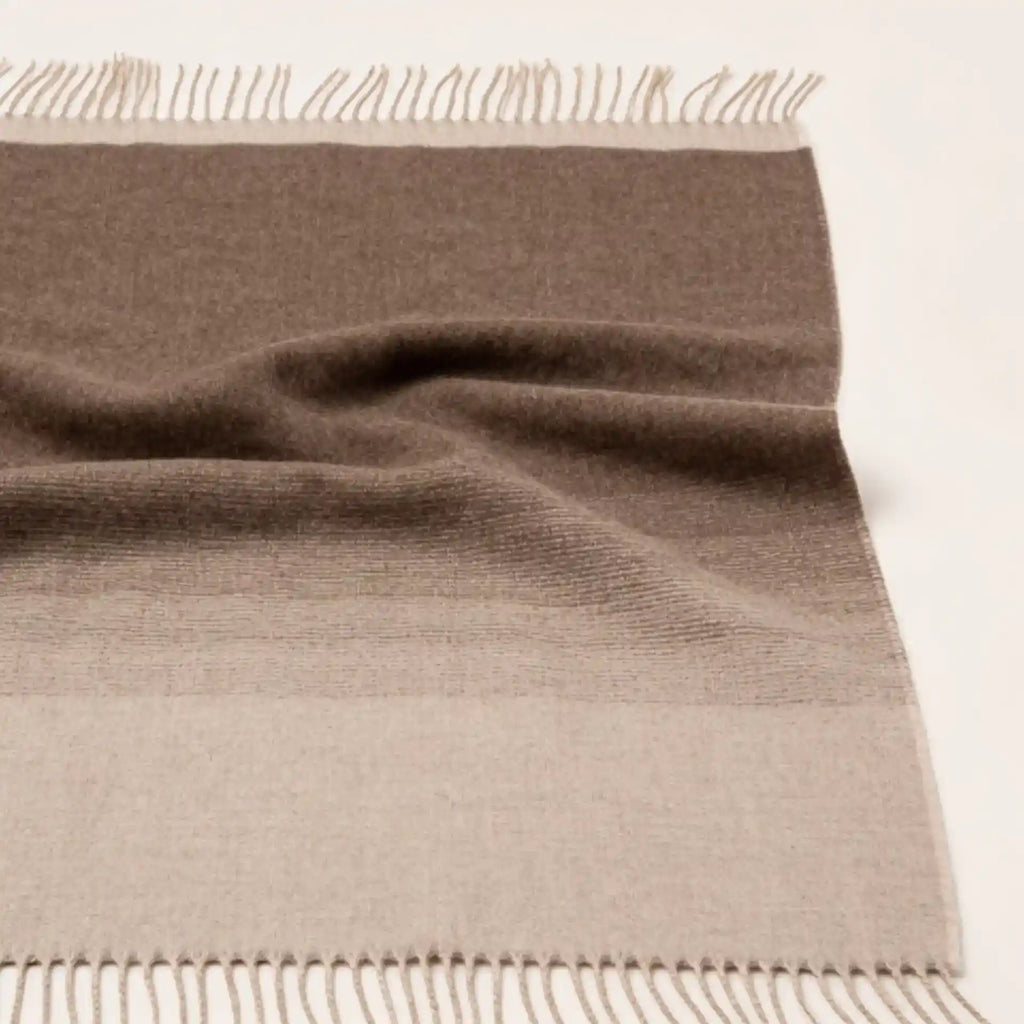 A folded woven throw blanket in taupe-brown and light beige tones with fringed edges, photographed on a plain light background.