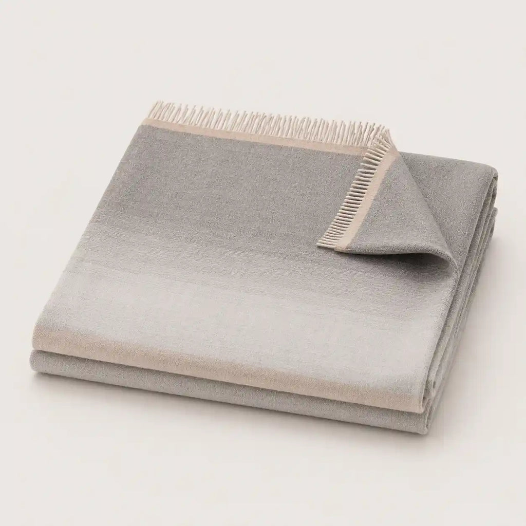 A folded light gray throw blanket with a subtle gradient and fringed edge, displayed on a clean off-white background.