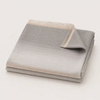 A folded light gray throw blanket with a subtle gradient and fringed edge, displayed on a clean off-white background.