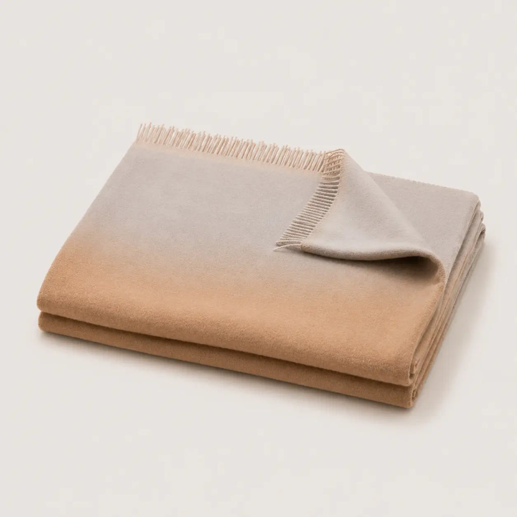A folded throw blanket with a smooth ombre gradient transitioning from warm tan to light gray, finished with fringed edges, shown on a clean off-white background.