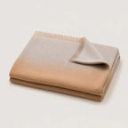 A folded throw blanket with a smooth ombre gradient transitioning from warm tan to light gray, finished with fringed edges, shown on a clean off-white background.
