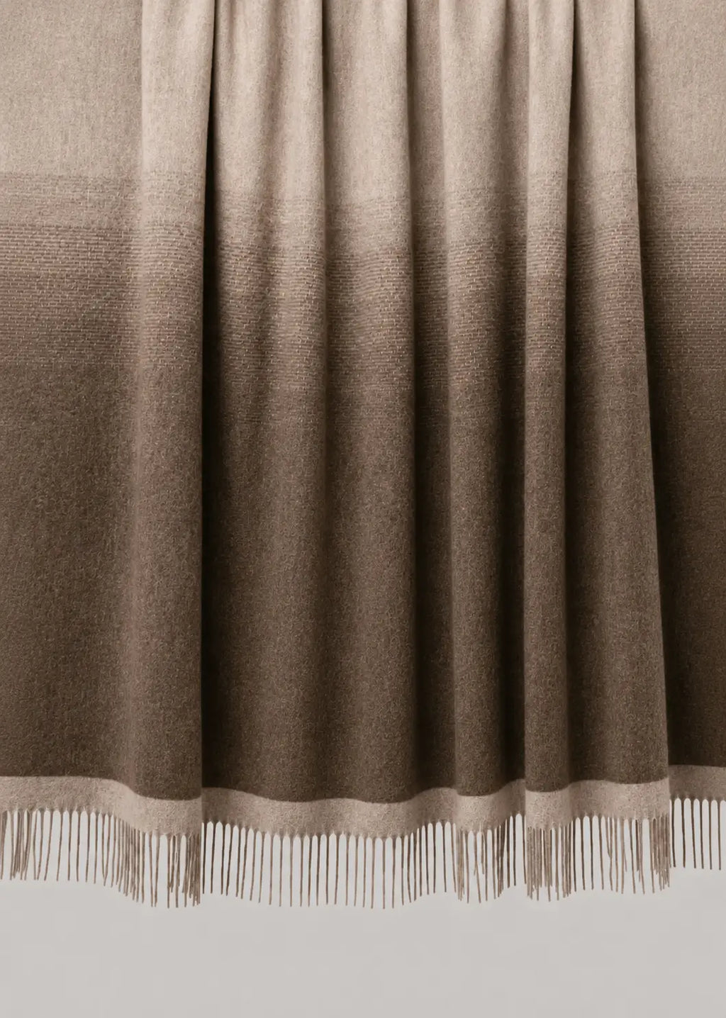 A woven throw blanket draped over a wooden rod, featuring a vertical ombre gradient from light beige at the top to dark brown at the bottom, finished with white fringe on the edge.