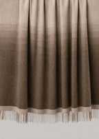 A woven throw blanket draped over a wooden rod, featuring a vertical ombre gradient from light beige at the top to dark brown at the bottom, finished with white fringe on the edge.