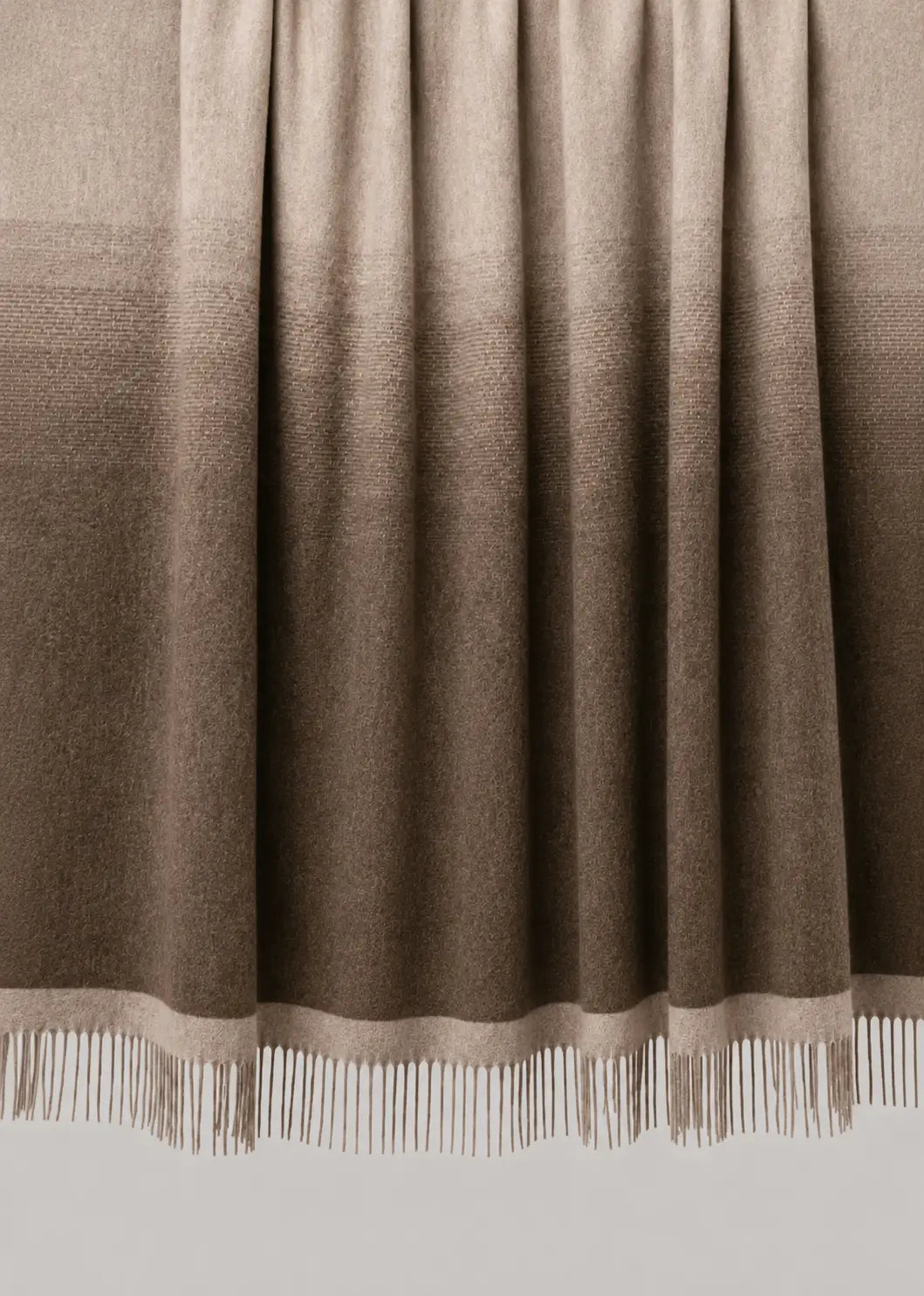 A woven throw blanket draped over a wooden rod, featuring a vertical ombre gradient from light beige at the top to dark brown at the bottom, finished with white fringe on the edge.