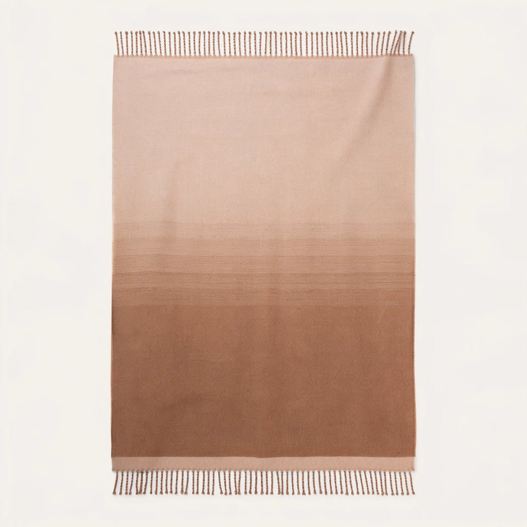 A rectangular woven throw blanket with a vertical ombré gradient transitioning from light beige at the top to a medium warm brown at the bottom, finished with fringed edges on a white background.