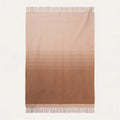 A rectangular woven throw blanket with a vertical ombré gradient transitioning from light beige at the top to a medium warm brown at the bottom, finished with fringed edges on a white background.