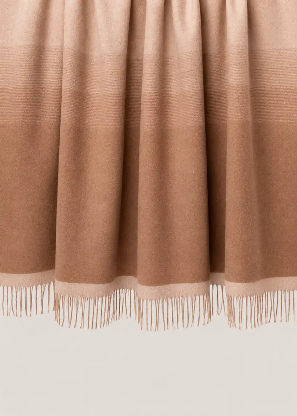 A close-up of a draped throw blanket or throw in a warm taupe-brown color, showing its fine texture and fringe along the bottom edge.