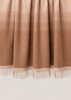 A close-up of a draped throw blanket or throw in a warm taupe-brown color, showing its fine texture and fringe along the bottom edge.