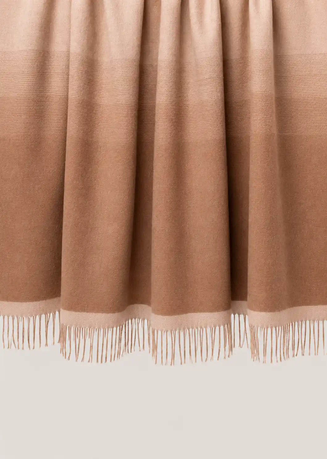 A close-up of a draped throw blanket or throw in a warm taupe-brown color, showing its fine texture and fringe along the bottom edge.