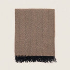 A rectangular woven throw blanket with a brown and beige herringbone pattern and black fringe along one edge, shown flat against a light off-white background.