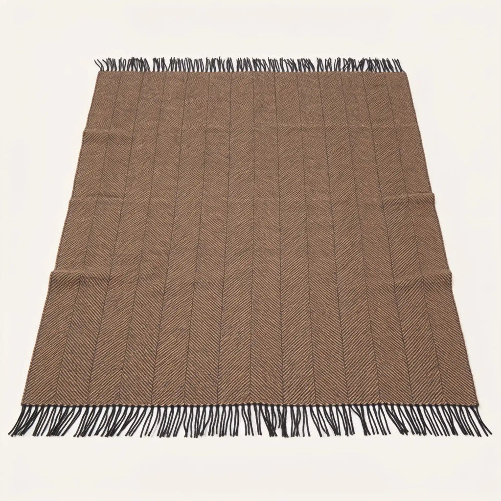A rectangular woven blanket throw in a warm taupe-brown colour with a subtle herringbone pattern and black fringe on all four ends, laid flat against a light off-white background.
