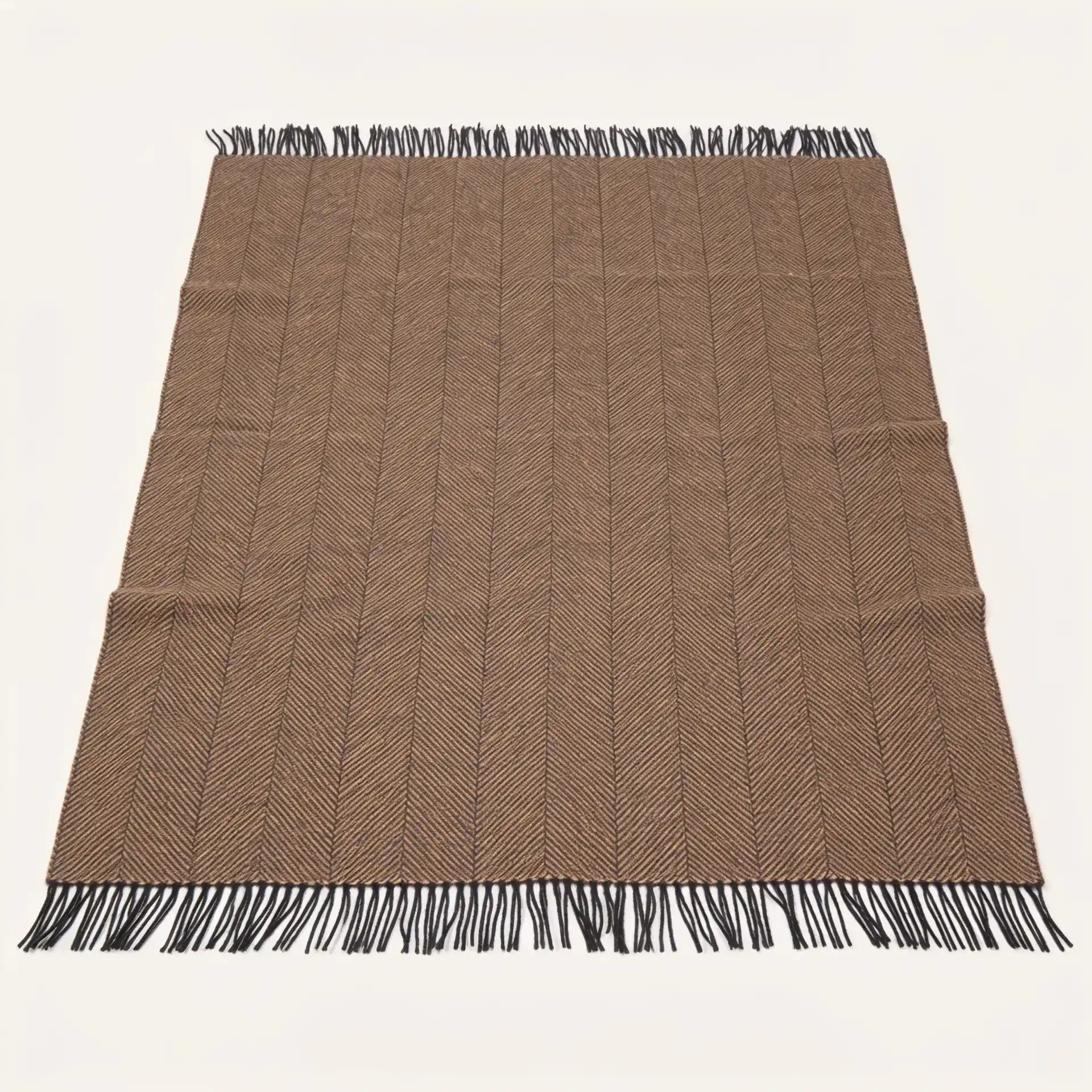 A rectangular woven blanket throw in a warm taupe-brown colour with a subtle herringbone pattern and black fringe on all four ends, laid flat against a light off-white background.