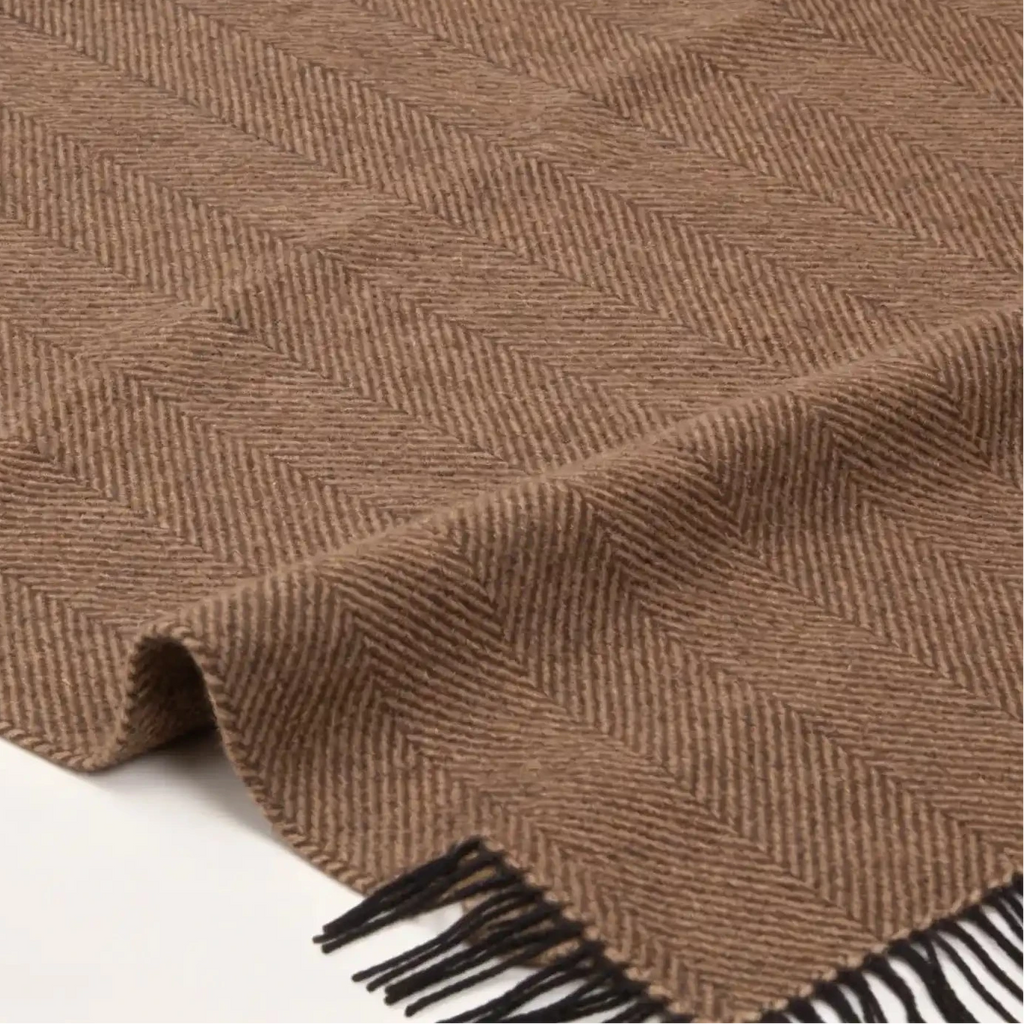 A brown woven blanket throw with a herringbone pattern and black fringe, laid out on a light background.