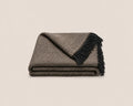 A folded woven throw blanket with a brown and beige herringbone pattern and black fringe, photographed on a light neutral background.