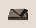 A folded woven throw blanket with a brown and beige herringbone pattern and black fringe, photographed on a light neutral background.