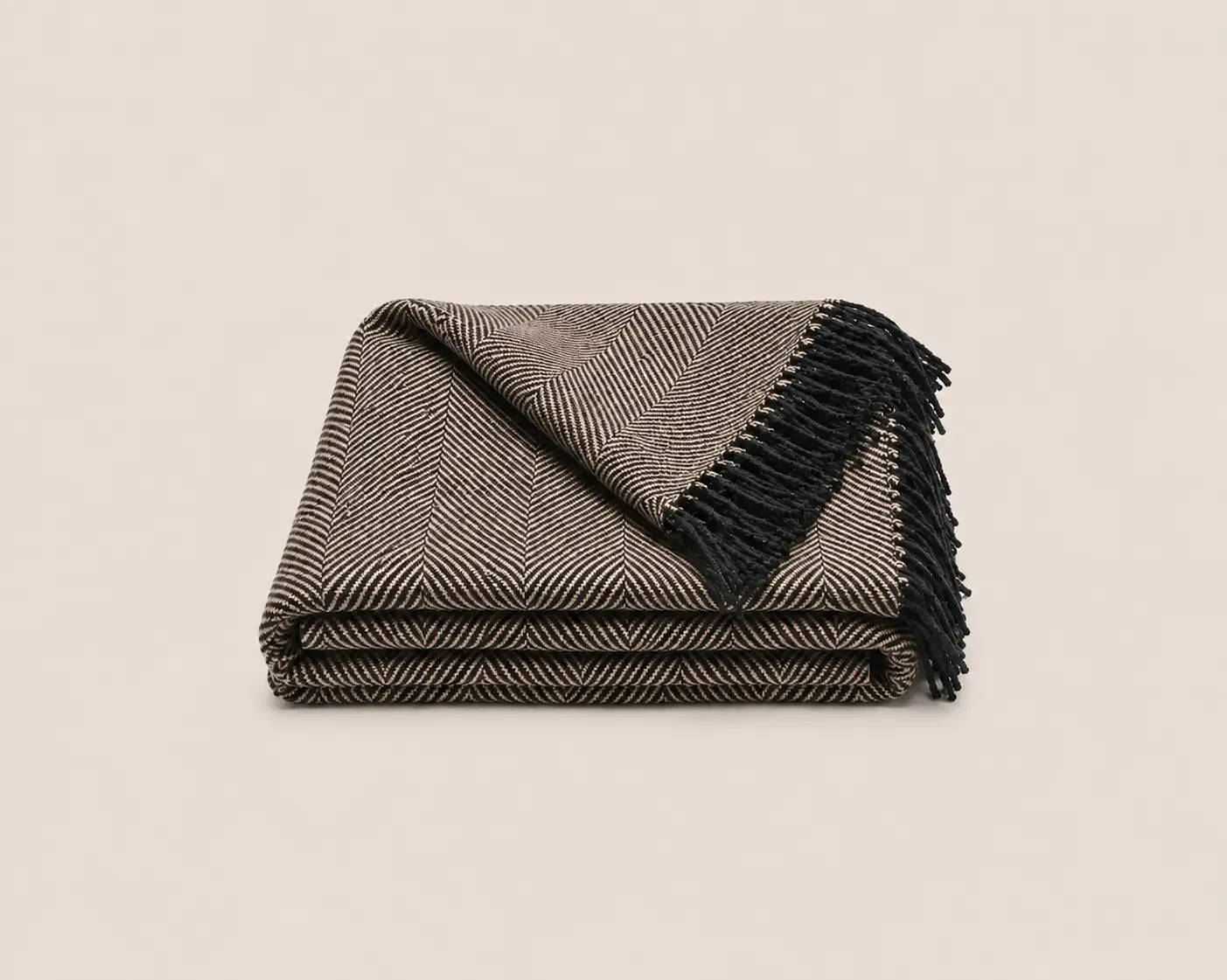 A folded woven throw blanket with a brown and beige herringbone pattern and black fringe, photographed on a light neutral background.