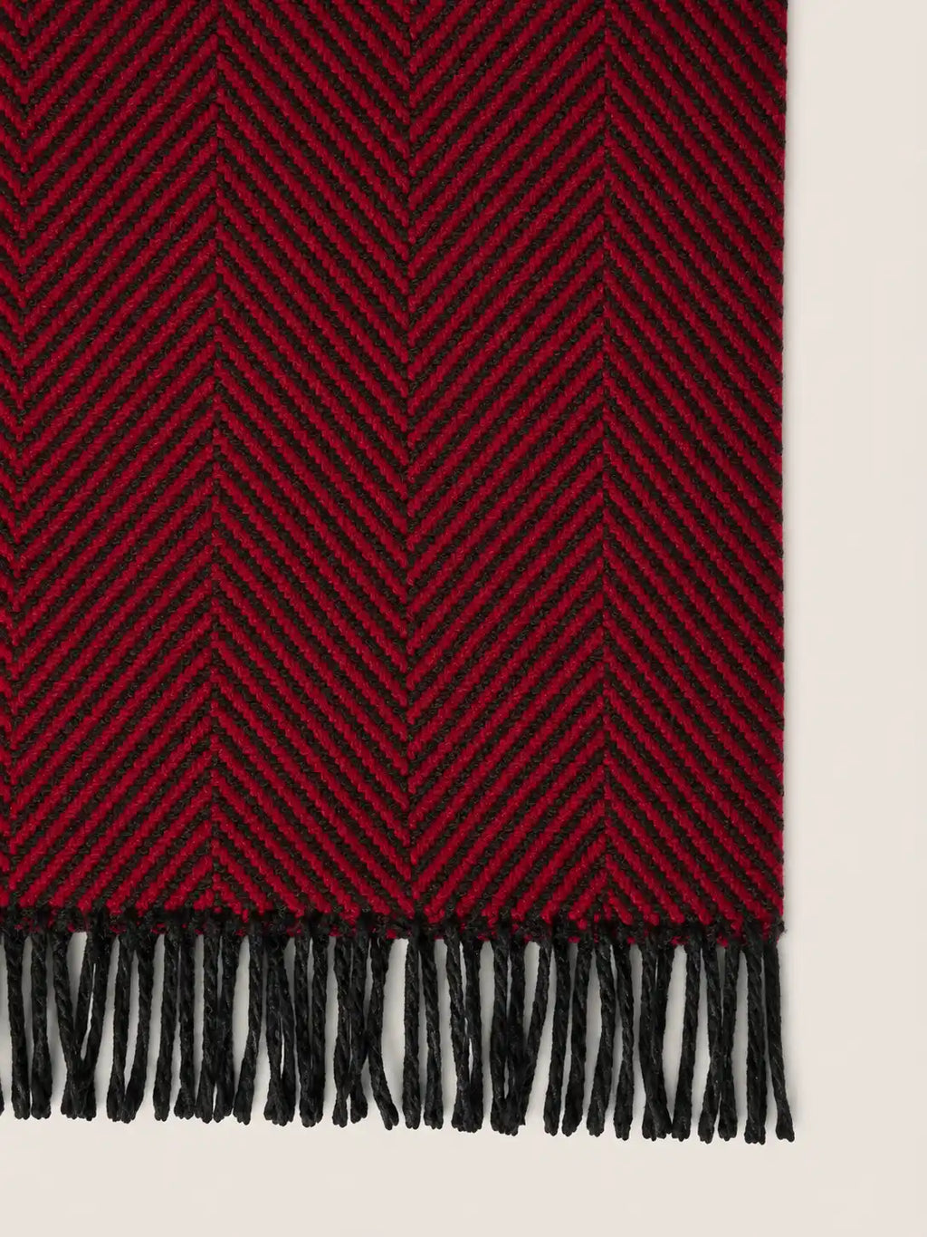 A close-up of a woven throw blanket with a deep red and black herringbone pattern and black fringe along one edge, shown against a light neutral background.