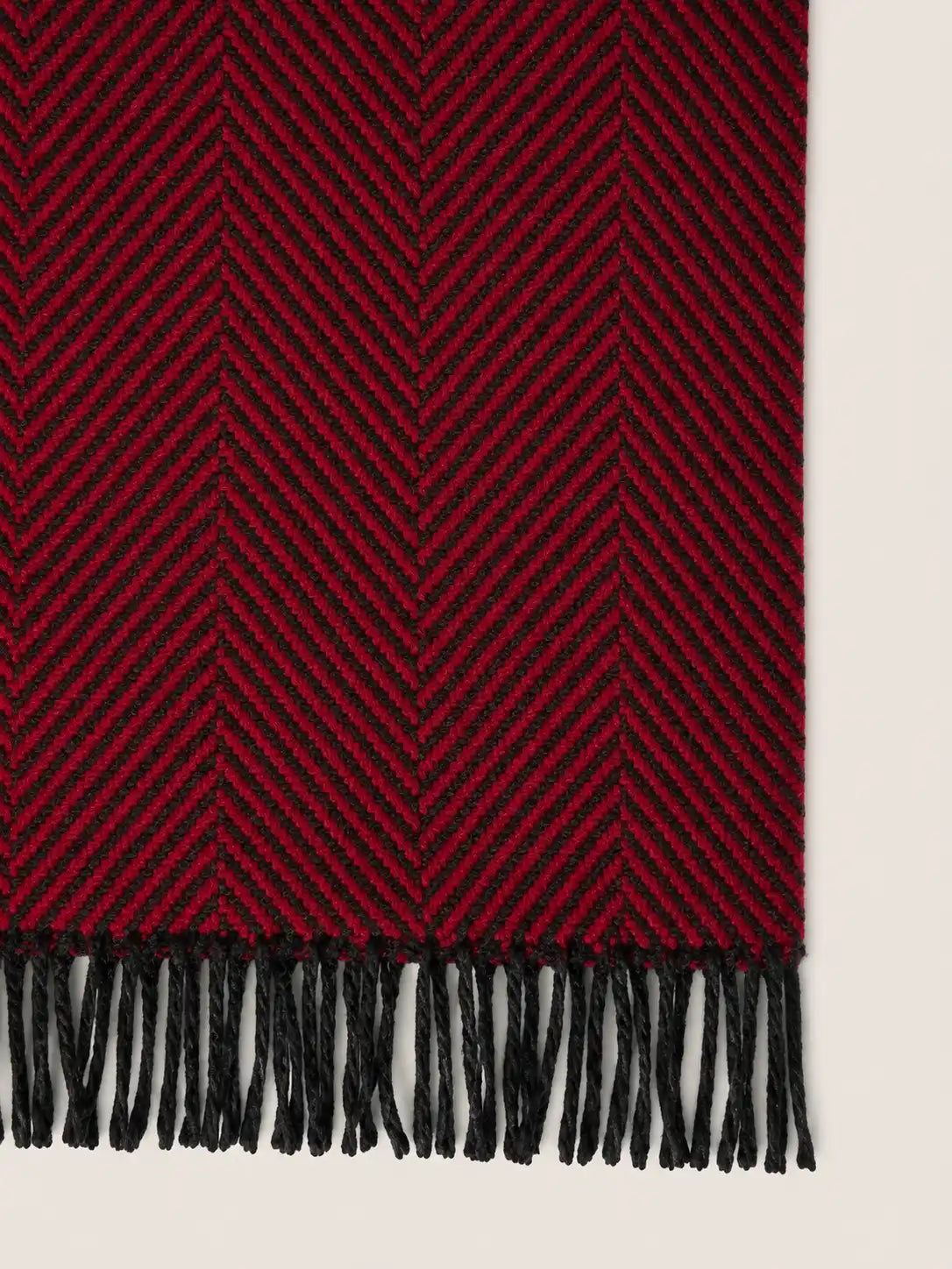 A close-up of a woven throw blanket with a deep red and black herringbone pattern and black fringe along one edge, shown against a light neutral background.