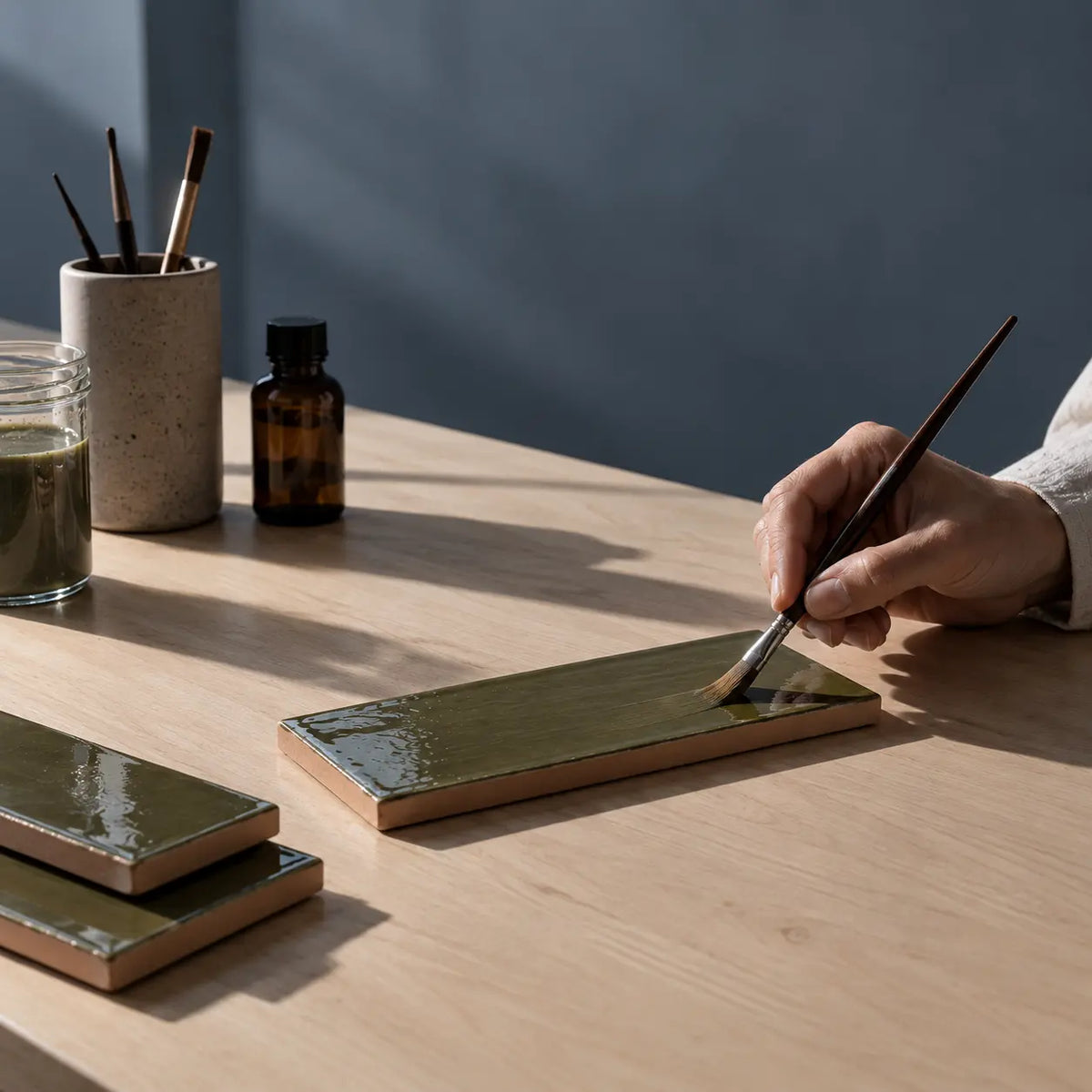 A hand holds a paintbrush applying glaze to a rectangular ceramic tile on a light wood table, with other glazed tiles and small bottles in the background.