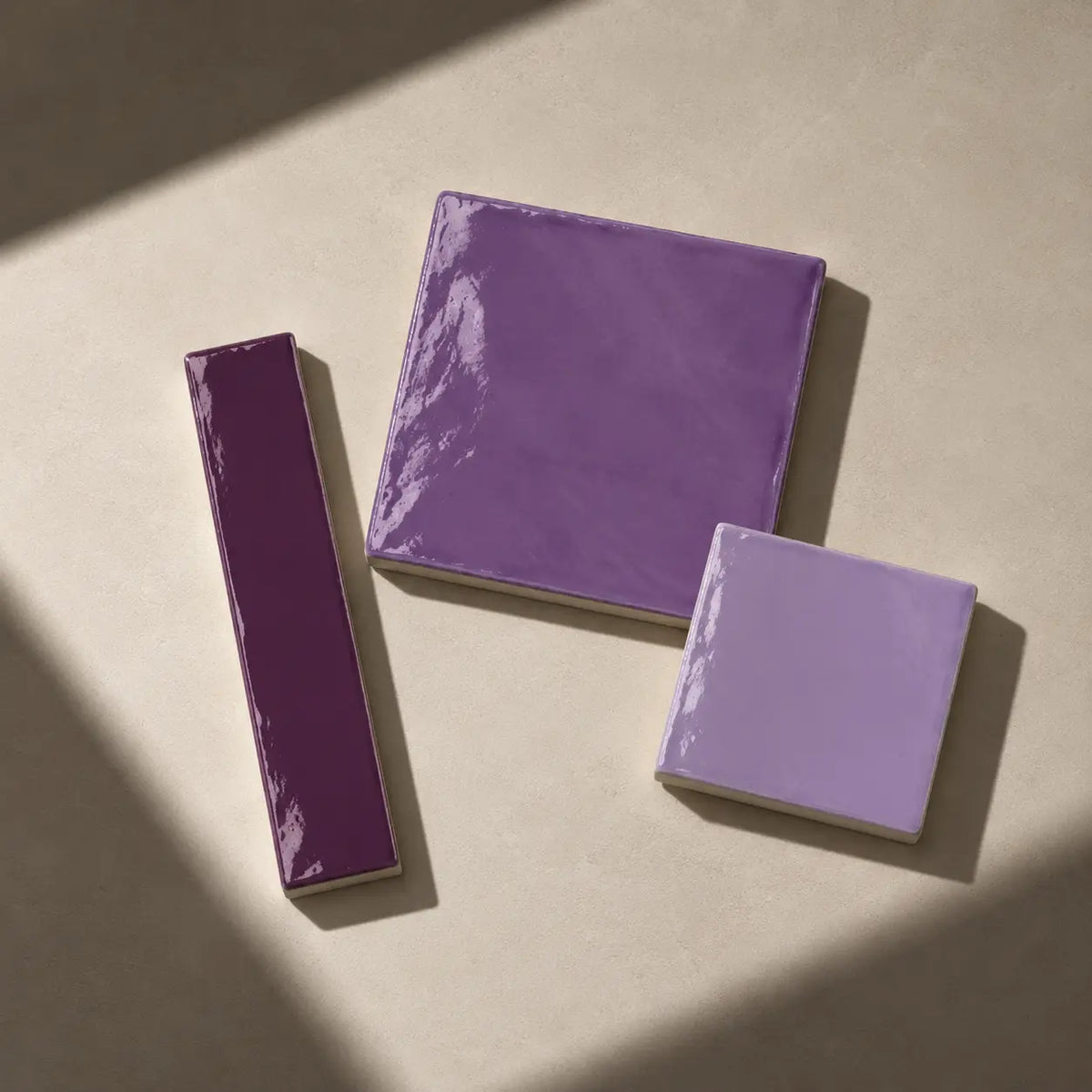 Three glossy purple color tiles in different shapes (one rectangular, two square) arranged on a textured beige surface with soft diagonal shadows.