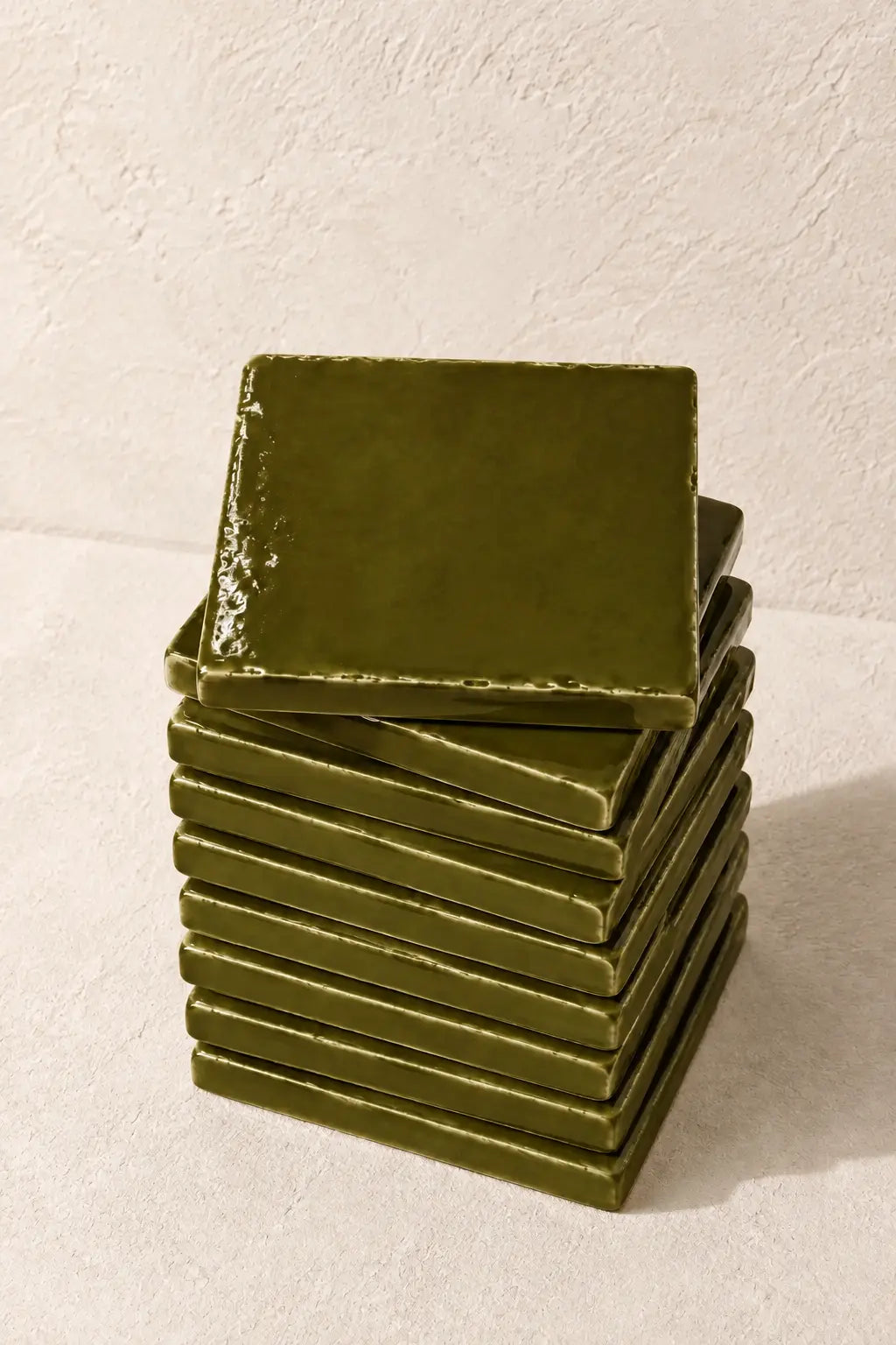 A stack of square moss green coasters with a glossy finish, arranged on a textured off-white surface against a matching background.