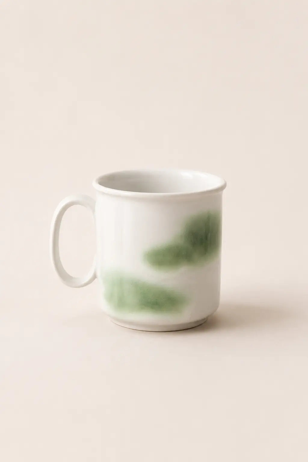 A white ceramic mug with a handle, decorated with soft green watercolor-like brushstrokes, set against a solid, warm off-white background.