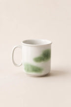 A white ceramic mug with a handle, decorated with soft green watercolor-like brushstrokes, set against a solid, warm off-white background.