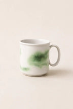 A white ceramic mug with a green watercolor-style wash on the side, set against a solid, warm off-white background.