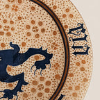 A round ceramic plate with a speckled beige and brown glaze, featuring a central dark blue dragon motif and Roman numerals around the rim, set against a plain light beige background.