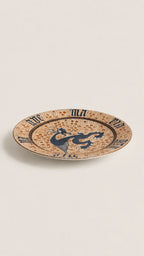 A round ceramic plate with a speckled beige and brown glaze, featuring a central dark blue dragon motif and Roman numerals around the rim, set against a plain light beige background.