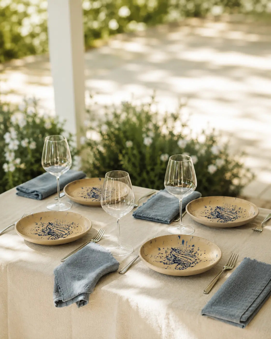 An outdoor dining table set with beige plates featuring a blue floral pattern, folded blue linen napkins, clear wine glasses, and gold-toned flatware under soft natural light.
