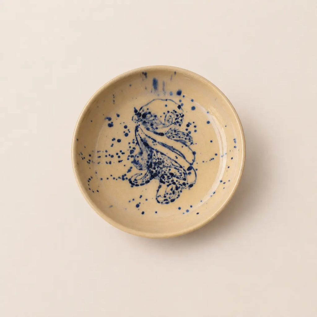 A round ceramic small plate with a speckled beige glaze and a hand-painted blue floral design in the center, photographed from directly above on a light beige background.