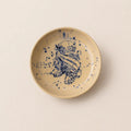 A round ceramic small plate with a speckled beige glaze and a hand-painted blue floral design in the center, photographed from directly above on a light beige background.