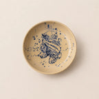 A round ceramic small plate with a speckled beige glaze and a hand-painted blue floral design in the center, photographed from directly above on a light beige background.